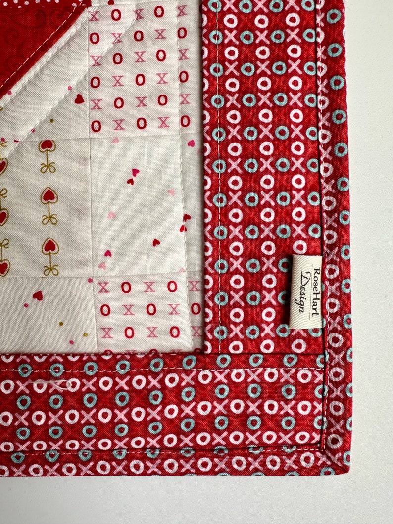 May include: A red, white, and blue fabric quilt with a heart pattern. The quilt is trimmed with a red fabric with a pattern of circles and Xs. The quilt has a label that reads "Rose Hart Design".