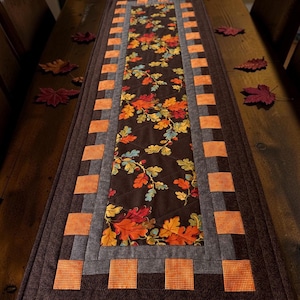May include: A rectangular table runner featuring a central panel with a dark brown background and a colourful pattern of autumn leaves in shades of orange, red, and yellow. The border consists of alternating dark brown and orange chequered squares.