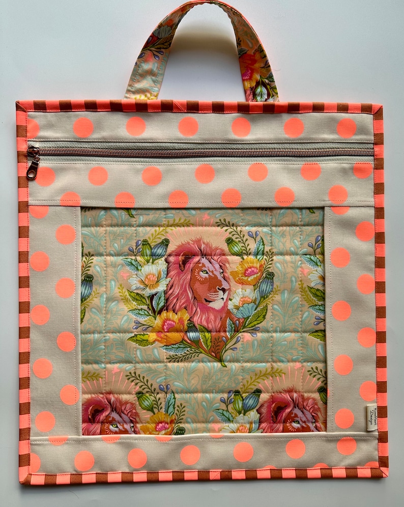 May include: A beige tote bag with a zipper closure and a large window featuring a quilted design with a lion and floral pattern. The bag has a brown and orange striped trim and pink polka dots.