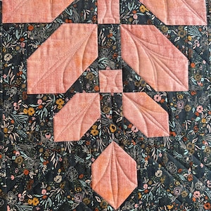 Pode incluir: Uma pe&ccedil;a de tecido acolchoado com fundo cinza escuro e uma estampa floral densa em tons de laranja, branco e azul-petr&oacute;leo. Formas de tecido s&oacute;lido na cor p&ecirc;ssego s&atilde;o aplicadas em um padr&atilde;o geom&eacute;trico com detalhes de costura vis&iacute;veis.