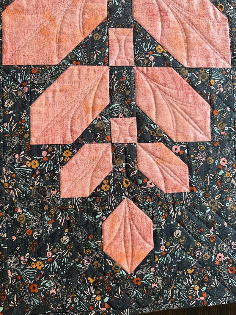 Pode incluir: Uma pe&ccedil;a de tecido acolchoado com fundo cinza escuro e uma estampa floral densa em tons de laranja, branco e azul-petr&oacute;leo. Formas de tecido s&oacute;lido na cor p&ecirc;ssego s&atilde;o aplicadas em um padr&atilde;o geom&eacute;trico com detalhes de costura vis&iacute;veis.