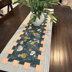 May include: A quilted table runner featuring a floral design in shades of green, yellow, and white. The runner has a border of peach-coloured gingham squares and a grey border with a subtle dotted pattern. The runner is displayed on a dark wood table with a vase of green olive branches.