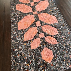Pode incluir: Um caminho de mesa acolchoado retangular com um design central de caule vertical com folhas de tecido na cor p&ecirc;ssego. O tecido de fundo &eacute; cinza escuro com uma estampa floral densa e multicolorida. A pe&ccedil;a inteira possui costuras acolchoadas em diagonal.