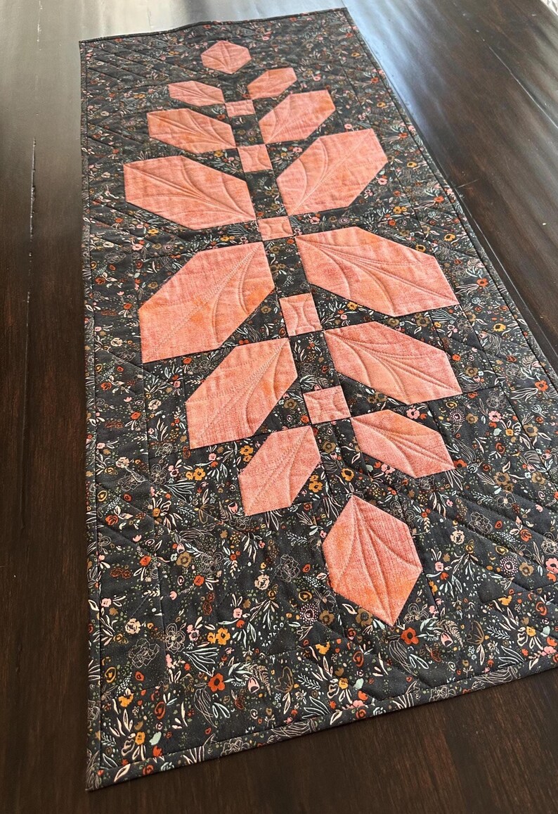 Pode incluir: Um caminho de mesa acolchoado retangular com um design central de caule vertical com folhas de tecido na cor p&ecirc;ssego. O tecido de fundo &eacute; cinza escuro com uma estampa floral densa e multicolorida. A pe&ccedil;a inteira possui costuras acolchoadas em diagonal.