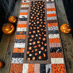 May include: A rectangular quilted table runner featuring a central panel with orange pumpkins on a black background, surrounded by a border of alternating fabric strips in orange, black, white, and gray with patterns of bats, spiders, and skeletons.