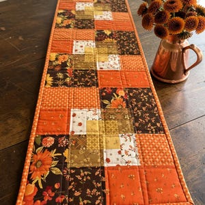 Puede incluir: Un camino de mesa acolchado rectangular con un diseño de patchwork de varias telas en tonos naranja, marrón, amarillo y crema. El patrón incluye estampados florales, pequeños motivos geométricos y cuadrados de color naranja sólido con costuras.