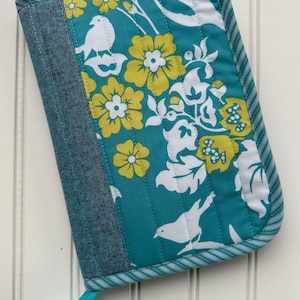 May include: A quilted book cover with a teal floral and bird pattern. The design features yellow flowers and white birds on a teal background. The spine is denim, and the edges are trimmed with striped fabric.