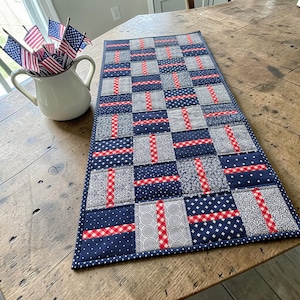 May include: A rectangular quilted table runner with a patriotic theme. The runner features a patchwork design with navy blue, red, and white fabrics. The design includes squares and rectangles with various patterns, including polka dots, stars, and gingham. A white pitcher with American flags is in the background.
