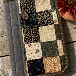May include: A rectangular quilted fabric cover featuring a patchwork design of black, cream, and olive green squares with floral and scroll patterns. A solid gray textured fabric panel runs along the left side, and the edges are finished with olive green binding.