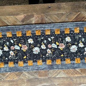 Puede incluir: Un camino de mesa gris y naranja con un patr&oacute;n floral. El camino de mesa tiene un fondo negro con flores blancas y amarillas. El camino de mesa est&aacute; acolchado y tiene un borde de cuadrados grises y naranjas.