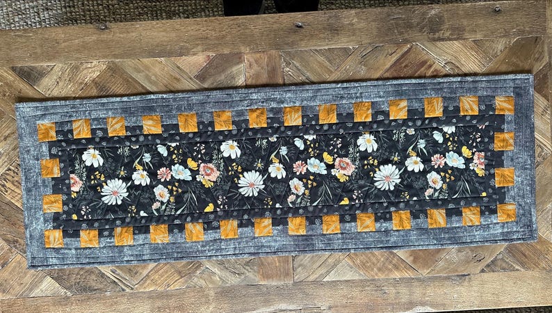 Puede incluir: Un camino de mesa gris y naranja con un patr&oacute;n floral. El camino de mesa tiene un fondo negro con flores blancas y amarillas. El camino de mesa est&aacute; acolchado y tiene un borde de cuadrados grises y naranjas.