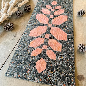 Pode incluir: Um caminho de mesa retangular acolchoado com um fundo de tecido cinza escuro com uma estampa floral densa em tons de laranja, branco e verde. Um design central vertical de folhas de tecido cor de p&ecirc;ssego est&aacute; costurado no caminho de mesa.