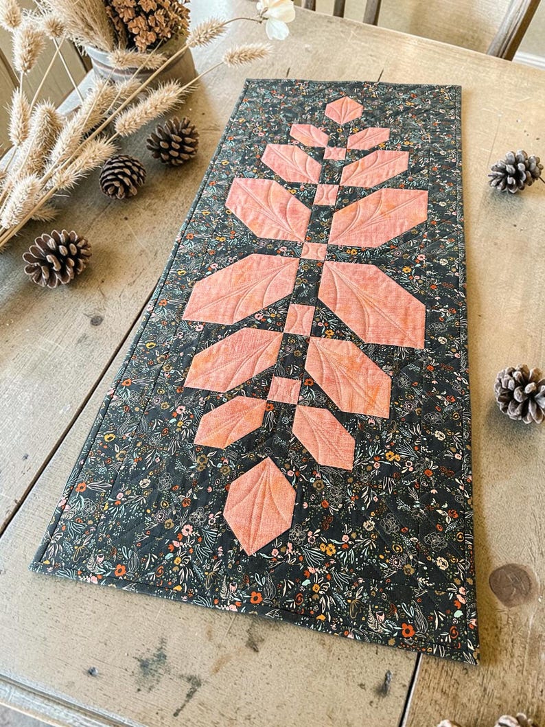 Pode incluir: Um caminho de mesa retangular acolchoado com um fundo de tecido cinza escuro com uma estampa floral densa em tons de laranja, branco e verde. Um design central vertical de folhas de tecido cor de p&ecirc;ssego est&aacute; costurado no caminho de mesa.
