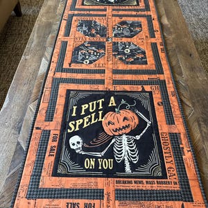 May include: Halloween-themed table runner with a patchwork design. The runner features orange, black, and gray fabrics with various spooky graphics and text, including a skeleton holding a pumpkin and the phrase "I PUT A SPELL ON YOU."