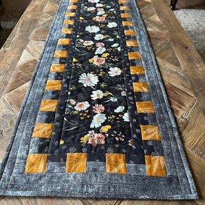 Puede incluir: Un camino de mesa floral blanco y negro con un borde amarillo mostaza. El camino de mesa tiene un dise&ntilde;o acolchado y es perfecto para agregar un toque de estilo farmhouse a tu mesa de comedor.