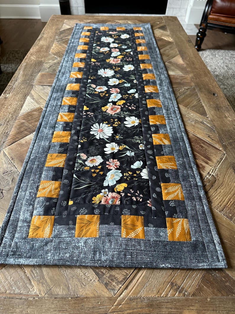 Puede incluir: Un camino de mesa floral blanco y negro con un borde amarillo mostaza. El camino de mesa tiene un dise&ntilde;o acolchado y es perfecto para agregar un toque de estilo farmhouse a tu mesa de comedor.