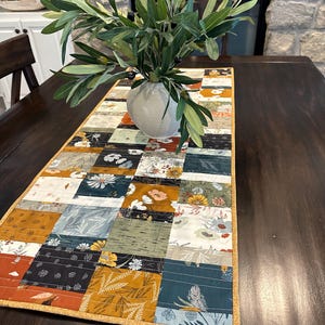 May include: A patchwork table runner with a floral and geometric design in shades of blue, green, yellow, and brown. The runner has a gold border and is displayed on a wooden table with a vase of olive branches.
