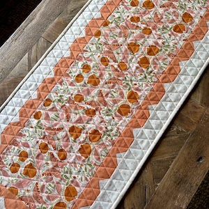 May include: A quilted table runner with a peach and white floral pattern. The runner has a scalloped edge and is made with a white fabric with a subtle pattern.