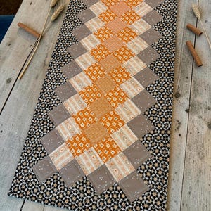 May include: A rectangular quilted table runner featuring a central diamond-shaped pattern of orange, cream, and taupe fabric squares. The border is made of black fabric with a small white floral print. The quilt is stitched with a grid design.