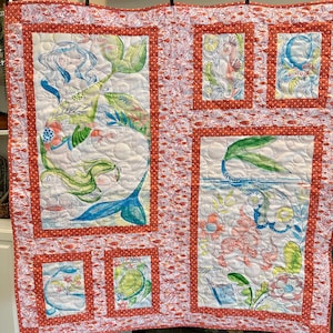 May include: A quilted wall hanging featuring mermaid illustrations. The quilt has a white background with colorful mermaid designs, framed by red borders and a patterned edge. The mermaid art includes blue, green, and coral colors.