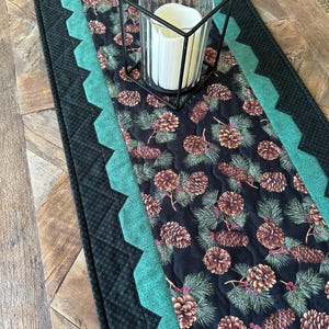 May include: A quilted table runner with a black background featuring a pine cone and pine needle pattern. The runner has a teal scalloped border and a black geometric patterned edge. A black metal candle holder with a white candle sits on top.