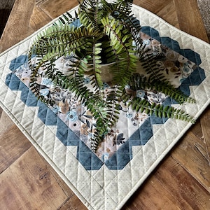 May include: A white quilted table runner with a blue and floral patterned center and a blue geometric border. A potted fern plant sits on top of the runner.