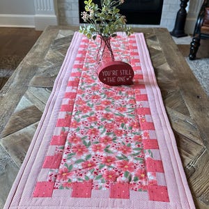 Puede incluir: Un camino de mesa acolchado con un estampado floral en tonos rosas y rojos. El camino de mesa tiene un borde rosa claro y un elemento decorativo que dice "YOU'RE STILL THE ONE". Un jarrón con vegetación encima.
