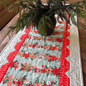 May include: A floral table runner with a light blue background and rows of red, orange, and peach flowers. The runner has a red scalloped edge and a green and white checkered border. A dark brown vase with green branches sits on top.