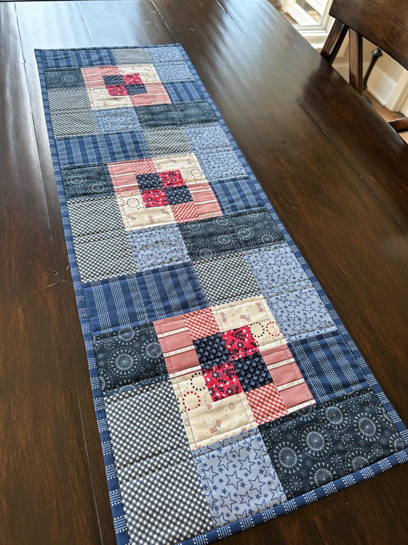Puede incluir: Un camino de mesa acolchado rectangular con un dise&ntilde;o de patchwork. El camino de mesa presenta cuadrados de varios patrones azules, incluyendo rayas, estrellas y motivos florales, intercalados con cuadrados estampados en rojo y blanco. Los bordes est&aacute;n acabados con un borde azul.