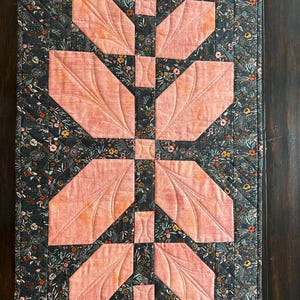 Pode incluir: Um caminho de mesa acolchoado retangular com um fundo de tecido cinza-carv&atilde;o e uma estampa floral densa em tons de laranja, branco e verde. O centro exibe um arranjo vertical de retalhos de tecido em forma de folha na cor p&ecirc;ssego.