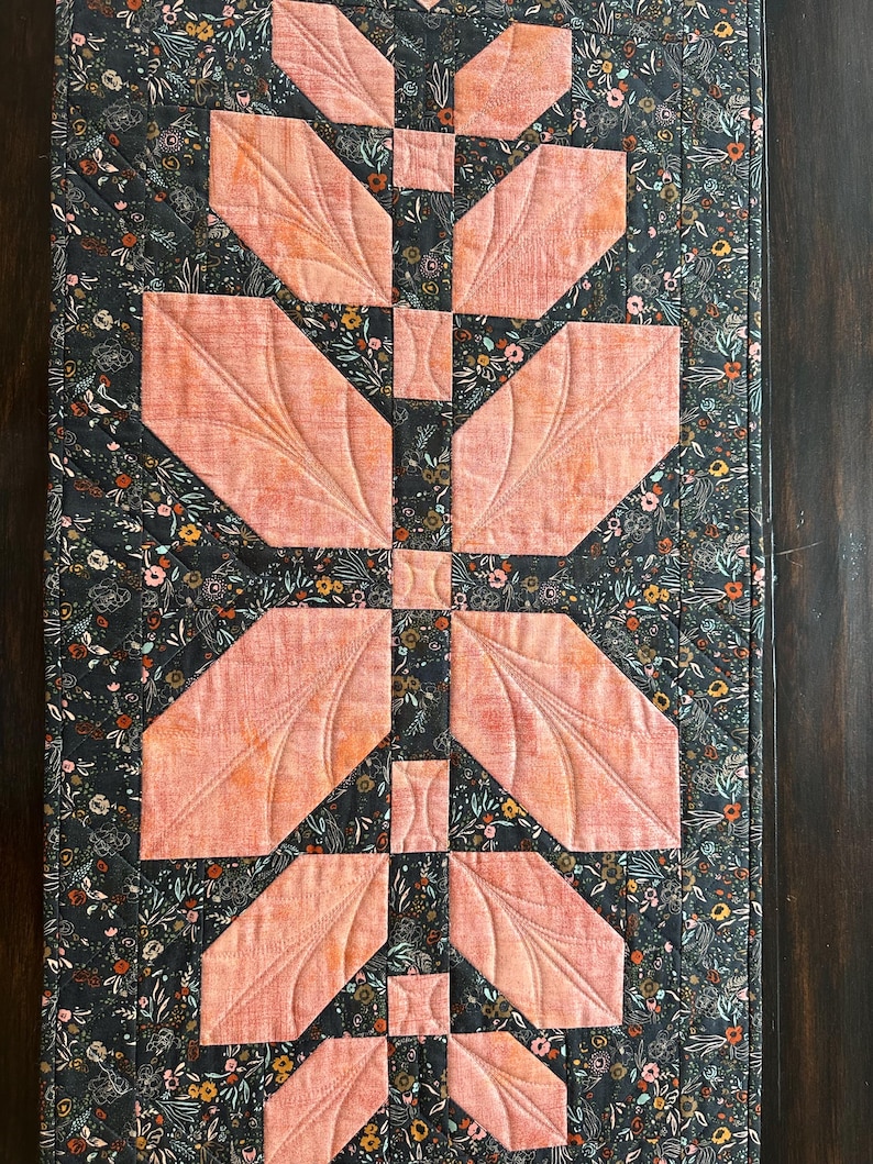 Pode incluir: Um caminho de mesa acolchoado retangular com um fundo de tecido cinza-carv&atilde;o e uma estampa floral densa em tons de laranja, branco e verde. O centro exibe um arranjo vertical de retalhos de tecido em forma de folha na cor p&ecirc;ssego.