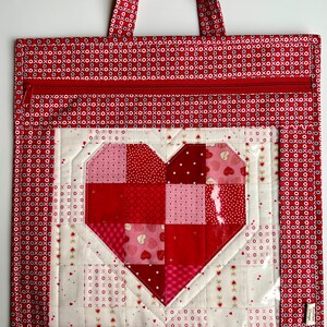 May include: Red fabric tote bag with a zipper closure and a clear vinyl window featuring a patchwork heart design. The heart is made of red, pink, and white fabric squares.