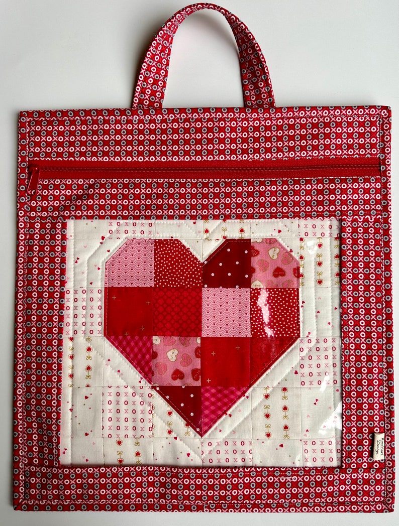 May include: Red fabric tote bag with a zipper closure and a clear vinyl window featuring a patchwork heart design. The heart is made of red, pink, and white fabric squares.