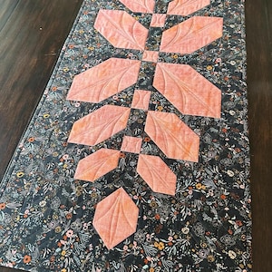 Pode incluir: Um caminho de mesa acolchoado retangular com um fundo de tecido cinza escuro com uma estampa floral densa em tons de laranja, branco e verde. Um design central vertical de folhas de tecido cor de p&ecirc;ssego &eacute; costurado em um padr&atilde;o sim&eacute;trico.