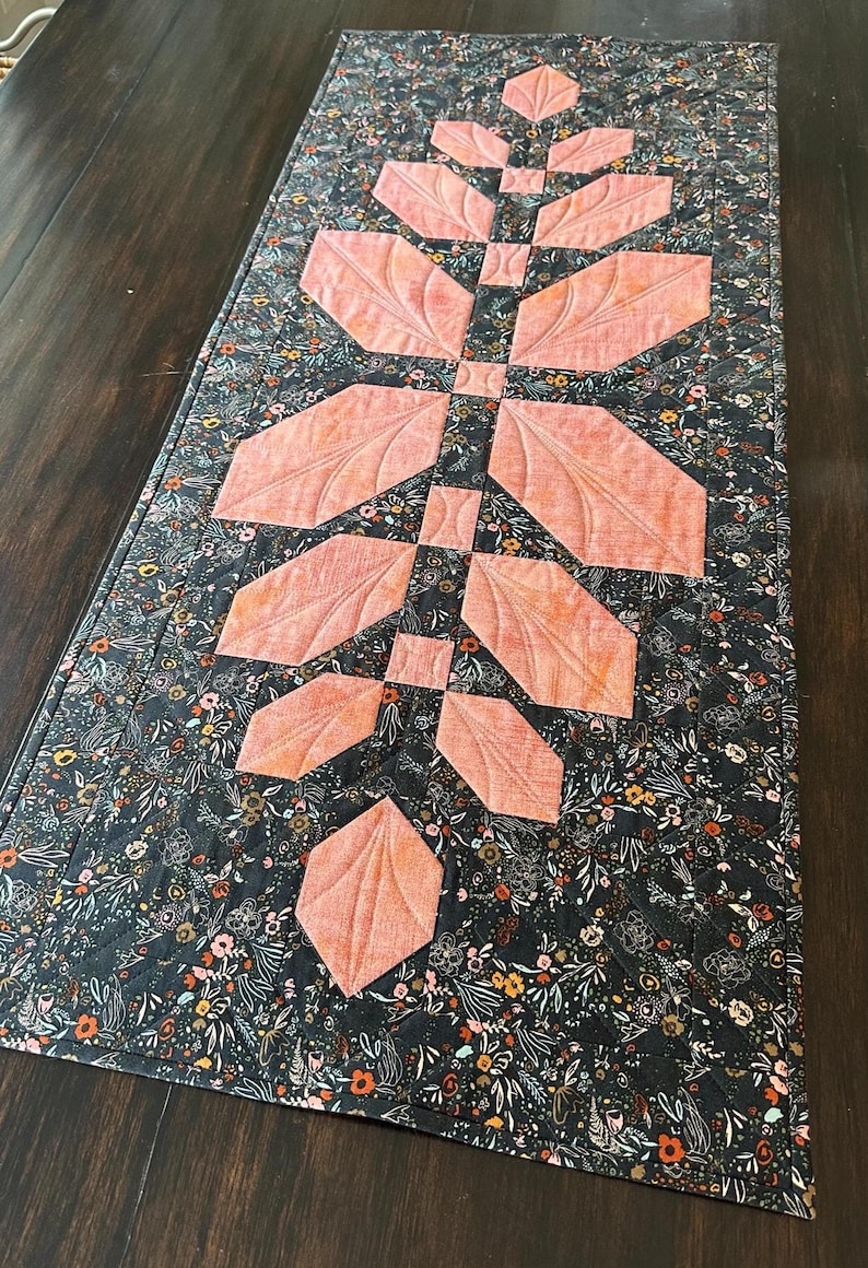 Pode incluir: Um caminho de mesa acolchoado retangular com um fundo de tecido cinza escuro com uma estampa floral densa em tons de laranja, branco e verde. Um design central vertical de folhas de tecido cor de p&ecirc;ssego &eacute; costurado em um padr&atilde;o sim&eacute;trico.