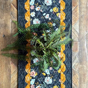 May include: A black quilted table runner with a floral print and a gold border. The runner is decorated with a repeating pattern of white and yellow flowers on a black background. The border is made of a gold fabric with a geometric pattern.