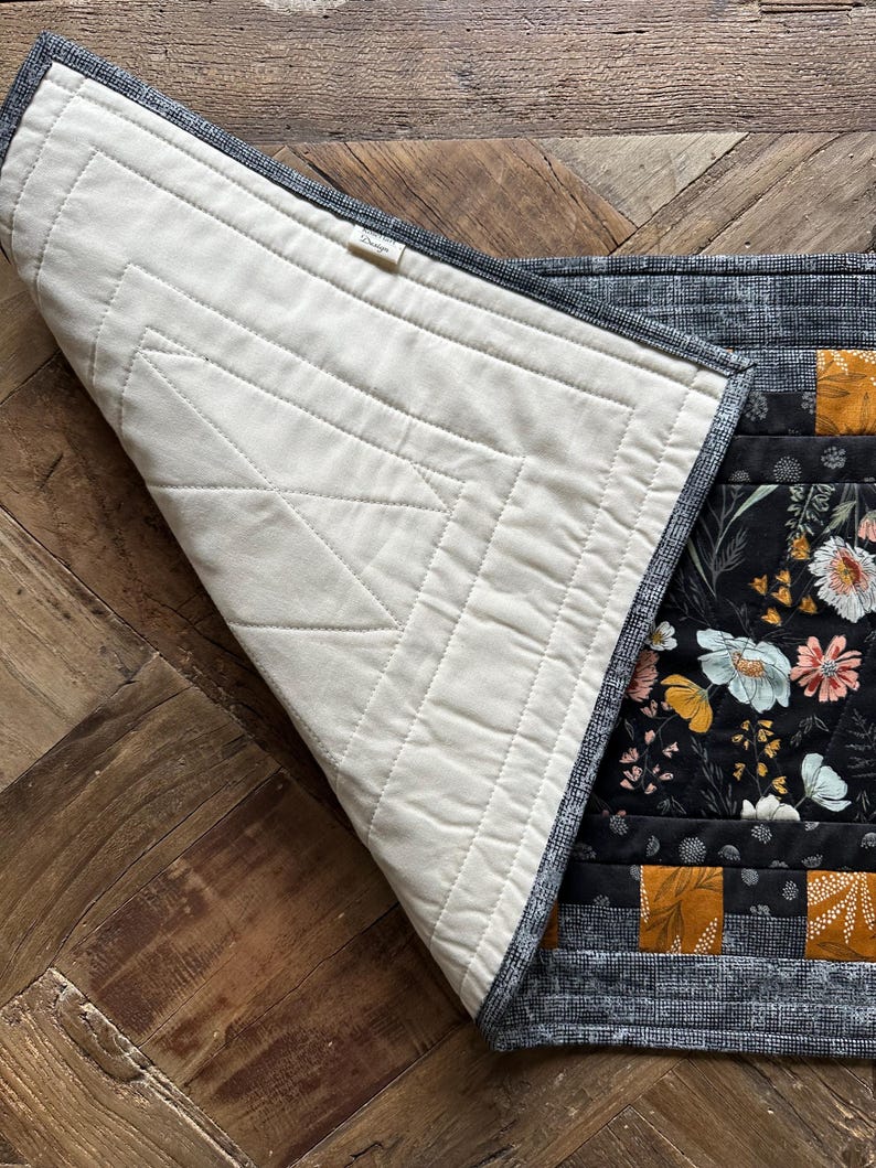 Puede incluir: Un camino de mesa de tela floral en blanco y negro con un patr&oacute;n geom&eacute;trico amarillo mostaza. El camino de mesa est&aacute; acolchado y tiene un respaldo de algod&oacute;n blanco. El camino de mesa est&aacute; doblado por la mitad y el respaldo es visible.