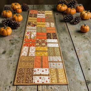 May include: A rectangular fabric table runner featuring a patchwork design of various autumn-themed patterns in shades of orange, yellow, brown, and cream. The runner is quilted with a thin border and displays motifs of leaves, flowers, and small pumpkins.