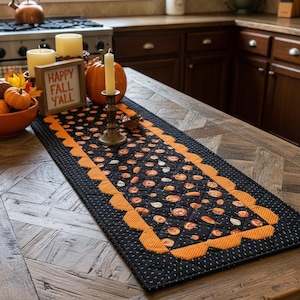May include: A quilted table runner featuring a black fabric with a pattern of small pumpkins and leaves, bordered by a scalloped orange edge. The runner is decorated with a wooden sign reading HAPPY FALL Y'ALL, a pumpkin, and two pillar candles.