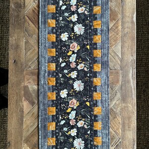 Puede incluir: Un camino de mesa floral blanco y negro con un borde amarillo y marr&oacute;n. El camino de mesa est&aacute; acolchado y tiene una textura sutil.