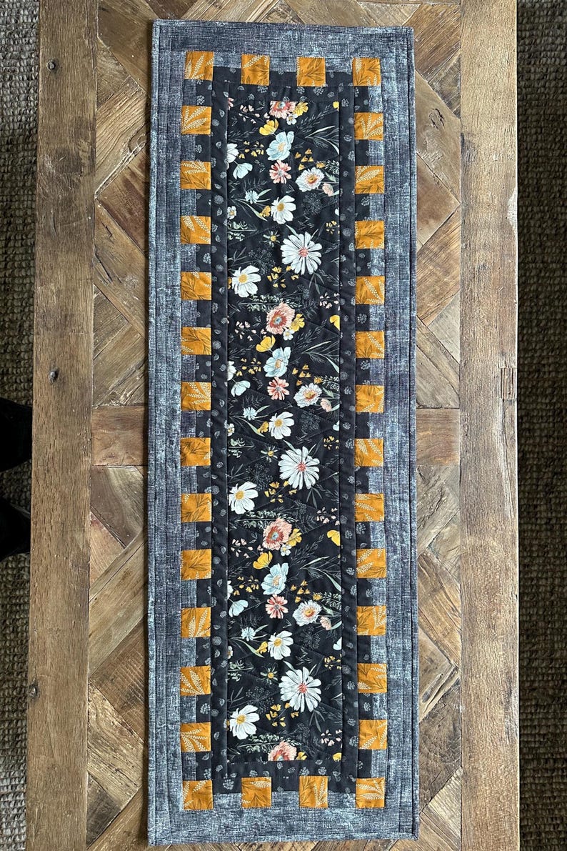Puede incluir: Un camino de mesa floral blanco y negro con un borde amarillo y marr&oacute;n. El camino de mesa est&aacute; acolchado y tiene una textura sutil.