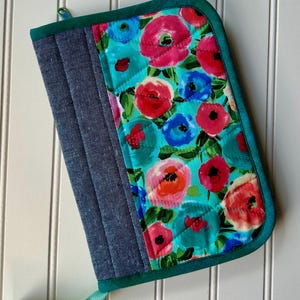 May include: Quilted oven mitt. One side is denim blue, the other features a floral pattern of pink, red, and blue flowers on a turquoise background. It has a green trim and a hanging loop.