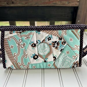 Peut inclure: Une trousse de maquillage matelassée avec un motif géométrique dans les tons de turquoise, marron et blanc. Le sac est orné d'une bordure à pois marron foncé et de poignées assorties. Le motif comprend des formes florales et abstraites.
