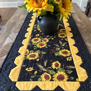 May include: A black quilted table runner with a sunflower pattern and yellow scalloped edges. A black vase holds a bouquet of yellow sunflowers. The runner is on a wooden table.
