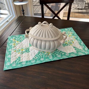 May include: A white ceramic serving dish with a lid, resting on a quilted table runner. The runner features a teal and white geometric pattern with floral accents. The dish has handles and a decorative finial on top.