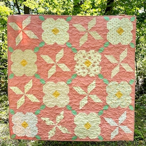 May include: A pink quilt with a repeating pattern of white flowers and green leaves. The quilt is made up of squares with different designs, including a starburst pattern and a floral pattern.