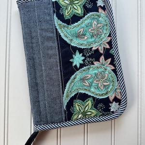 May include: A quilted case with a denim-like fabric on the left and a paisley pattern on the right. The paisley design features teal, pink, and green floral elements on a navy blue background. The case is trimmed with a striped border.
