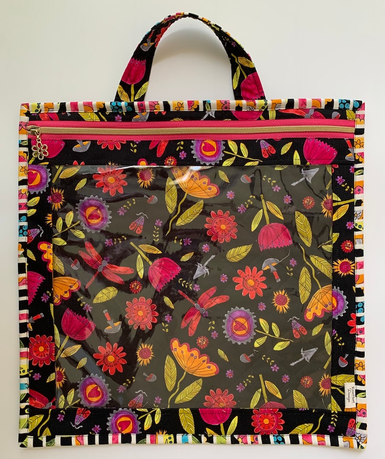 May include: Black zippered tote bag with a clear vinyl window. The bag has a floral print with pink, yellow, and red flowers and dragonflies on a black background. The bag has a pink zipper and a gold zipper pull.