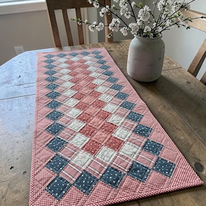 May include: A quilted table runner with a diamond pattern in shades of red, white, and blue. The runner features a gingham border and is placed on a wooden table. A vase of white flowers sits nearby.