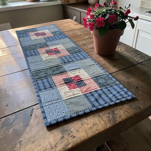 Puede incluir: Camino de mesa rectangular hecho a mano con dise&ntilde;o de patchwork. Presenta cuadrados de telas estampadas en azul, rosa y blanco. Se encuentra sobre una mesa de madera r&uacute;stica. Una maceta con flores rojas est&aacute; cerca. Mide aproximadamente 100 cm de largo.